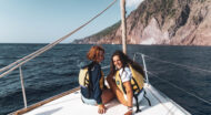 Two women sitting on the back of a sailboat in the ocean.